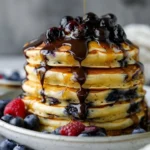 Blueberry pancakes topped with chocolate ganache and berry sauce on a plate.